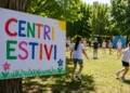 Bambini partecipano ad attività all'aperto durante un centro estivo in un parco, con un cartello colorato "Centri Estivi" in primo piano.