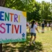 Bambini partecipano ad attività all'aperto durante un centro estivo in un parco, con un cartello colorato "Centri Estivi" in primo piano.