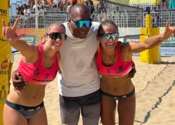 Due atlete di beach volley in divisa rosa festeggiano insieme a un uomo al centro del campo da gioco, davanti agli spalti con il pubblico