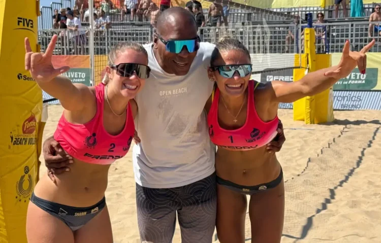 Due atlete di beach volley in divisa rosa festeggiano insieme a un uomo al centro del campo da gioco, davanti agli spalti con il pubblico