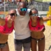 Due atlete di beach volley in divisa rosa festeggiano insieme a un uomo al centro del campo da gioco, davanti agli spalti con il pubblico