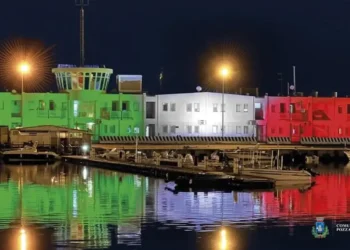 La sede della Capitaneria di Porto di Pozzallo illuminata con i colori del tricolore italiano riflessi sul mare di notte.
