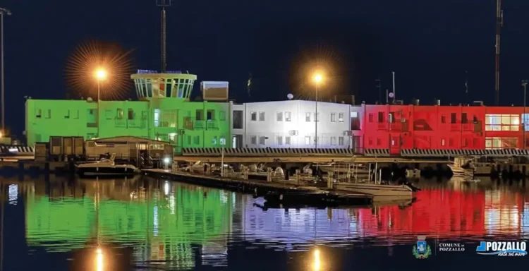 La sede della Capitaneria di Porto di Pozzallo illuminata con i colori del tricolore italiano riflessi sul mare di notte.