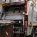 Operatore ecologico in gilet giallo-verde carica un bidone marrone su un camion della raccolta rifiuti in una via del centro storico