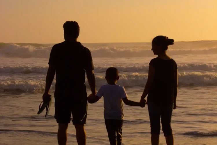 Una famiglia con un bambino osserva il mare al tramonto, tenendosi per mano sulla battigia. Immagine simbolica dedicata al sostegno economico per le famiglie e all'assegno unico.