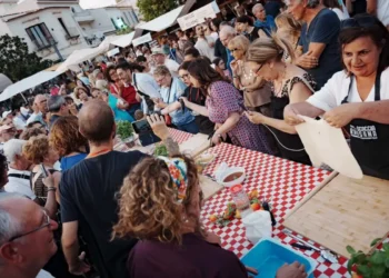 Pubblico numeroso assiste a una dimostrazione di preparazione della scaccia durante la Festa della Scaccia, con cuoche al lavoro attorno a un lungo tavolo nel centro storico di Ragusa.