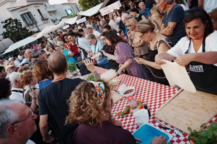 Pubblico numeroso assiste a una dimostrazione di preparazione della scaccia durante la Festa della Scaccia, con cuoche al lavoro attorno a un lungo tavolo nel centro storico di Ragusa.