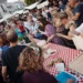 Pubblico numeroso assiste a una dimostrazione di preparazione della scaccia durante la Festa della Scaccia, con cuoche al lavoro attorno a un lungo tavolo nel centro storico di Ragusa.