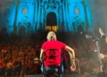 Cantautore di spalle sul palco con chitarra, folla sulla scalinata e facciata barocca della chiesa di San Giorgio illuminata di blu, Modica