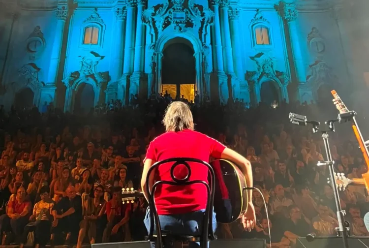 Cantautore di spalle sul palco con chitarra, folla sulla scalinata e facciata barocca della chiesa di San Giorgio illuminata di blu, Modica
