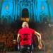 Cantautore di spalle sul palco con chitarra, folla sulla scalinata e facciata barocca della chiesa di San Giorgio illuminata di blu, Modica