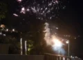 Fotografia notturna che mostra l'inizio di un incendio sulla collina del Belvedere a Modica, illuminato dai fuochi d'artificio e da fumo, con lampioni stradali e vegetazione in primo piano