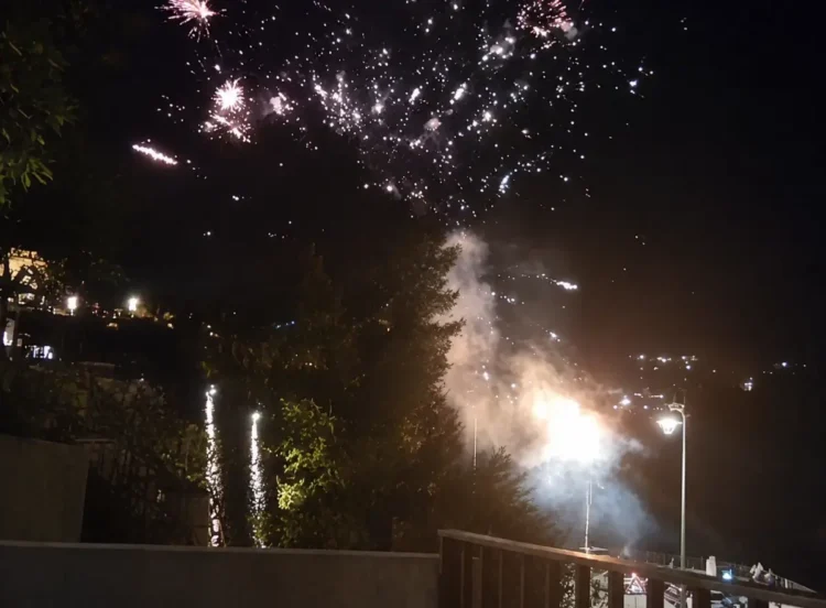 Fotografia notturna che mostra l'inizio di un incendio sulla collina del Belvedere a Modica, illuminato dai fuochi d'artificio e da fumo, con lampioni stradali e vegetazione in primo piano