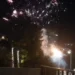 Fotografia notturna che mostra l'inizio di un incendio sulla collina del Belvedere a Modica, illuminato dai fuochi d'artificio e da fumo, con lampioni stradali e vegetazione in primo piano