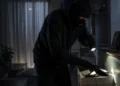 Figura incappucciata con passamontagna e guanti neri rovista in un cassetto al buio tenendo una torcia elettrica