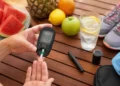 Persona che misura la glicemia con un glucometro accanto a frutta fresca, un bicchiere d'acqua con limone e scarpe da ginnastica, simboli di idratazione, alimentazione sana e attività fisica durante l'estate.