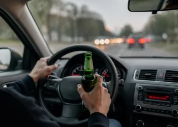Mani al volante di un'auto in marcia: la sinistra regge una bottiglia di birra verde, la destra impugna il volante