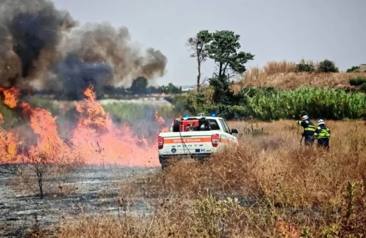Vigili del fuoco e Protezione Civile impegnati nello spegnimento di un vasto incendio di sterpaglie a Santa Croce Camerina, con alte fiamme e una densa colonna di fumo.