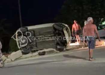 Auto ribaltata su un fianco dopo un incidente stradale a Sampieri, con alcuni residenti presenti sul luogo dell'impatto