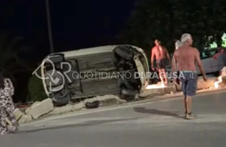 Auto ribaltata su un fianco dopo un incidente stradale a Sampieri, con alcuni residenti presenti sul luogo dell'impatto