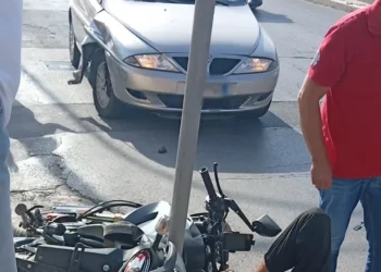 Moto a terra dopo uno scontro con un'auto grigia in strada a Vittoria; un uomo in rosso osserva la scena