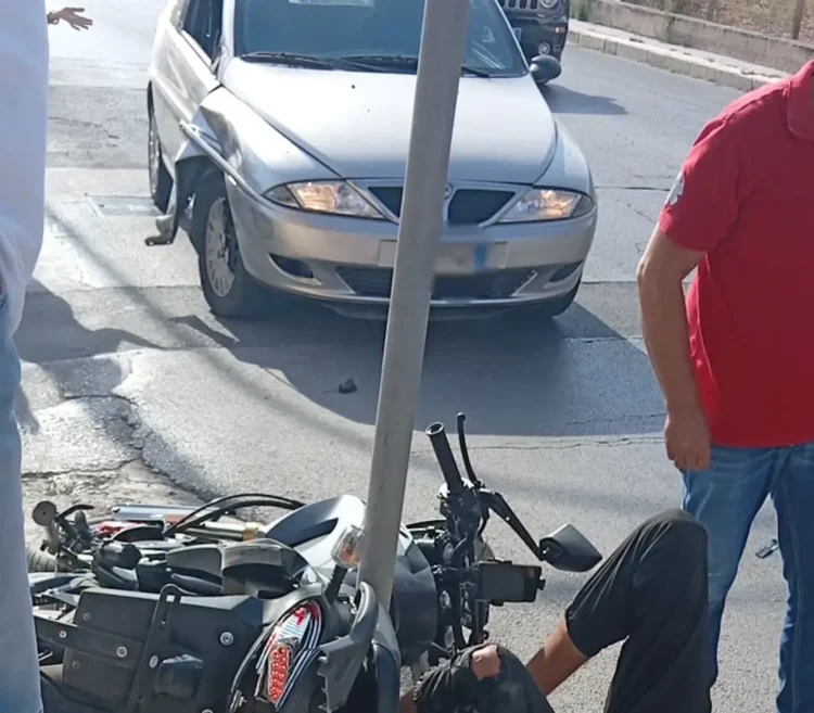 Moto a terra dopo uno scontro con un'auto grigia in strada a Vittoria; un uomo in rosso osserva la scena