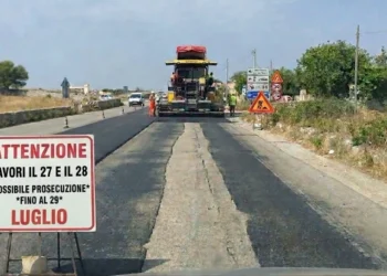 Macchina asfaltrice al lavoro sulla Sp 25 Ragusa-Marina con cartello che annuncia lavori il 27 e 28 luglio, possibile prosecuzione al 29