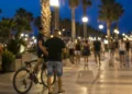 Persone passeggiano sul lungomare di Marina di Ragusa nelle ore serali, mentre un ciclista conduce la bicicletta a mano nell'area pedonale.
