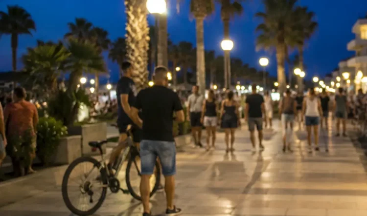 Persone passeggiano sul lungomare di Marina di Ragusa nelle ore serali, mentre un ciclista conduce la bicicletta a mano nell'area pedonale.