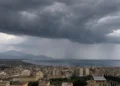 Panoramica di una città siciliana affacciata sul mare con un imponente fronte temporalesco, cielo coperto da nubi scure e piogge visibili in lontananza