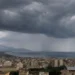 Panoramica di una città siciliana affacciata sul mare con un imponente fronte temporalesco, cielo coperto da nubi scure e piogge visibili in lontananza