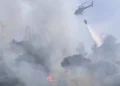 Elicottero antincendio impegnato nelle operazioni di spegnimento di un vasto incendio boschivo in Sicilia, con una benna d'acqua sopra un'area avvolta da fumo e fiamme
