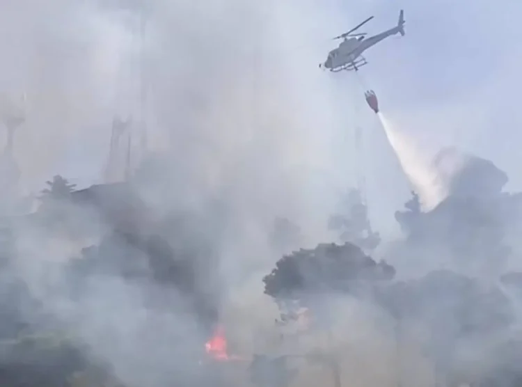 Elicottero antincendio impegnato nelle operazioni di spegnimento di un vasto incendio boschivo in Sicilia, con una benna d'acqua sopra un'area avvolta da fumo e fiamme