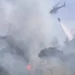 Elicottero antincendio impegnato nelle operazioni di spegnimento di un vasto incendio boschivo in Sicilia, con una benna d'acqua sopra un'area avvolta da fumo e fiamme