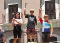 Tre giovani ciclisti sul podio della quarta tappa di ciclismo su strada a Palagonia, con i trofei alzati al termine della gara. Al centro un atleta della Motyka Bike School Modica.