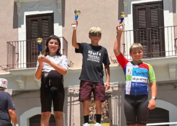 Tre giovani ciclisti sul podio della quarta tappa di ciclismo su strada a Palagonia, con i trofei alzati al termine della gara. Al centro un atleta della Motyka Bike School Modica.