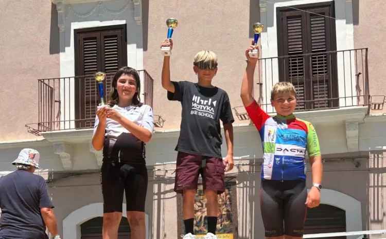 Tre giovani ciclisti sul podio della quarta tappa di ciclismo su strada a Palagonia, con i trofei alzati al termine della gara. Al centro un atleta della Motyka Bike School Modica.