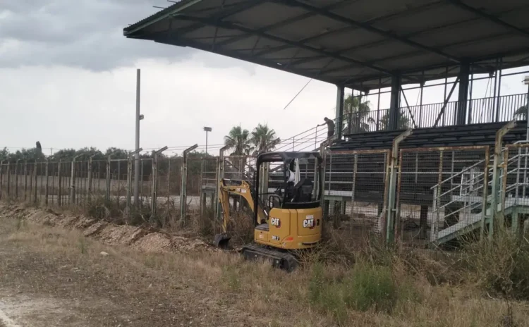 Mini escavatore CAT al lavoro lungo il perimetro del campo sportivo di Pedalino, accanto alla tribuna metallica