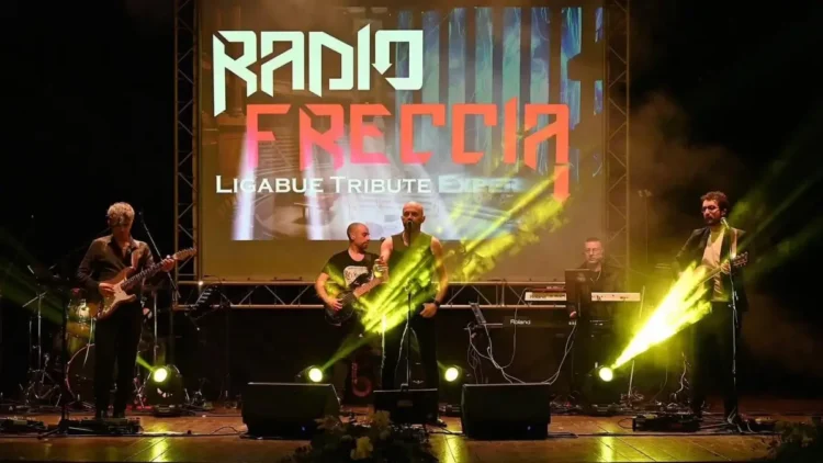 I Radiofreccia sul palco con luci gialle e schermo con il logo della band, durante un concerto all'aperto.
