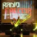 I Radiofreccia sul palco con luci gialle e schermo con il logo della band, durante un concerto all'aperto.