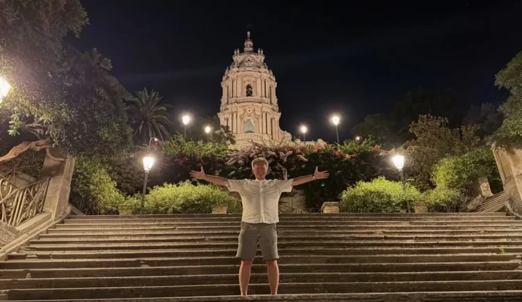 Red Ronnie posa con le braccia aperte sulla scalinata del Duomo di San Giorgio a Modica, illuminato di sera durante la sua vacanza in provincia di Ragusa