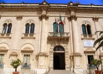 Facciata del Municipio di Scicli con colonne, balcone con bandiere italiana, europea e siciliana, ingresso ad arco
