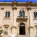 Facciata del Municipio di Scicli con colonne, balcone con bandiere italiana, europea e siciliana, ingresso ad arco