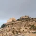 Panoramica di Scicli con la Chiesa di San Matteo sul colle che domina il centro storico, tra abitazioni in pietra e cielo nuvoloso.