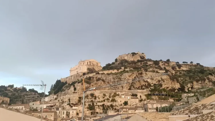 Veduta panoramica del colle di San Matteo a Scicli con l'antica chiesa sulla sommità e il centro storico ai piedi della collina sotto un cielo nuvoloso