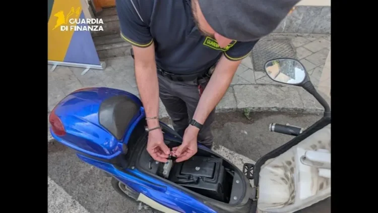 Un finanziere esamina i componenti elettrici interni di uno scooter blu durante un'operazione di sequestro