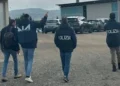Agenti DIA e Polizia di spalle durante un'operazione in un'area industriale con capannoni e parcheggio.