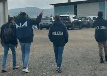 Agenti DIA e Polizia di spalle durante un'operazione in un'area industriale con capannoni e parcheggio.