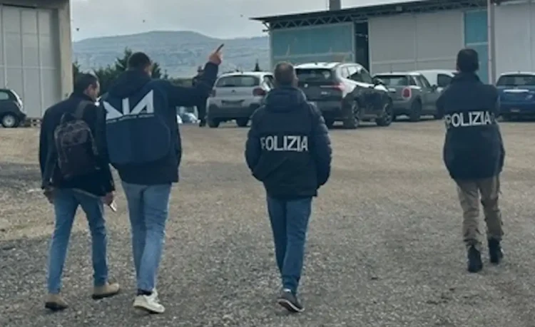 Agenti DIA e Polizia di spalle durante un'operazione in un'area industriale con capannoni e parcheggio.