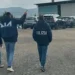Agenti DIA e Polizia di spalle durante un'operazione in un'area industriale con capannoni e parcheggio.
